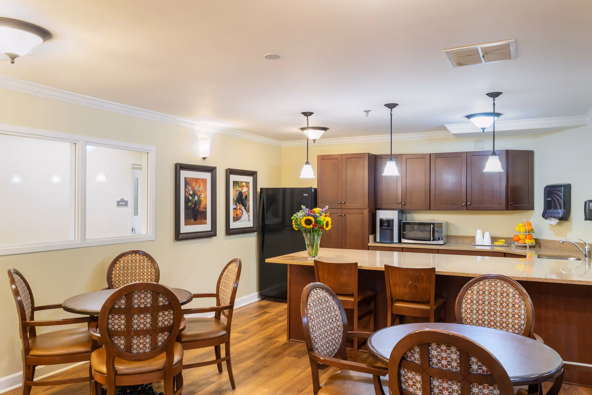 Communal dining area with round tables and chairs facing a kitchenette and breakfast bar topped with a vase of sunflowers.