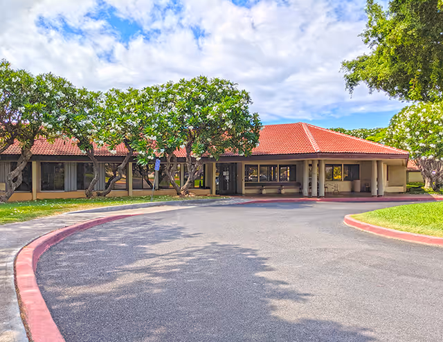 A single-story health facility with a red-tiled roof, curved driveway, and shaded entrance.