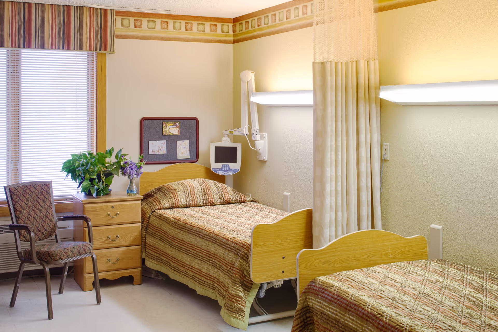 A room with two single beds with wooden headboards and patterned bedspreads. Between the beds is a wooden nightstand with a potted plant and a vase of purple flowers. A chair with patterned upholstery is next to the nightstand. A window with blinds and a striped valance is on the left wall. A medical monitor is mounted on the wall above one bed, and a privacy curtain hangs between the beds. The walls are light-colored with a decorative border near the ceiling.