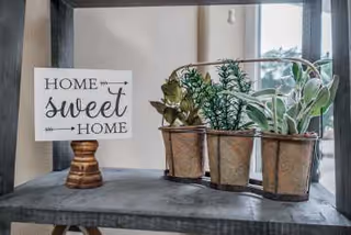 A decorative wooden sign that reads 'Home sweet Home' placed on a small wooden stand next to three small potted green plants on a gray shelf near a window.