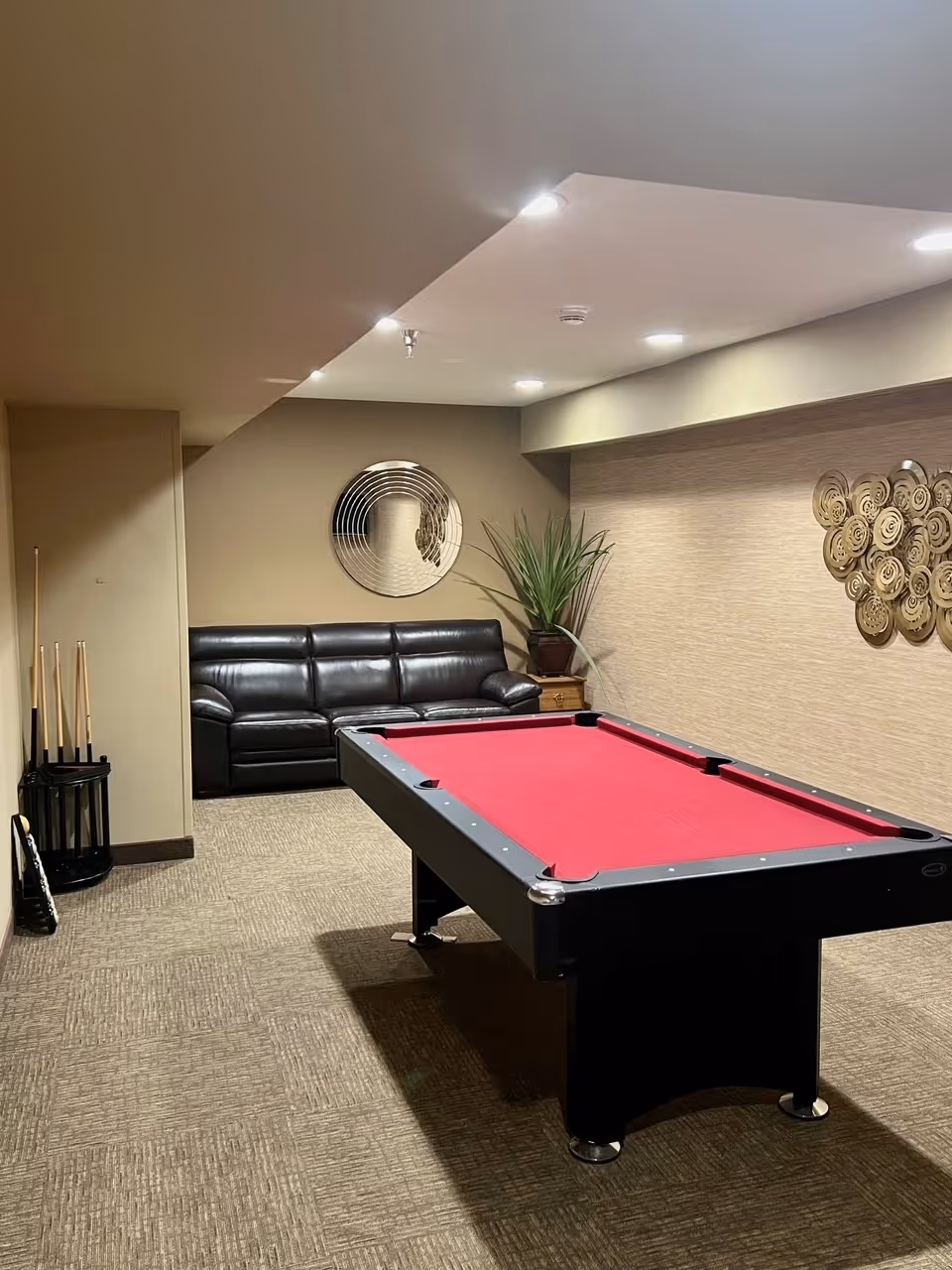 A small recreation room featuring a red-felt pool table, black leather sofa, wall art, a potted plant, and a rack of pool cues.