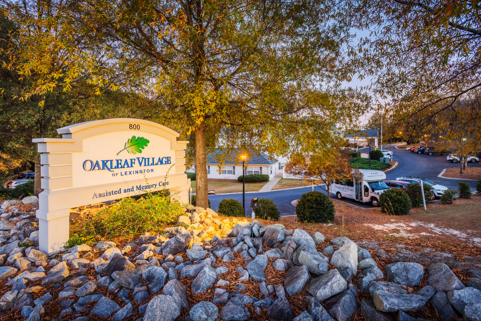 Entrance sign for Oakleaf Village of Lexington on a landscaped rocky slope with the facility buildings and a shuttle bus visible behind it.