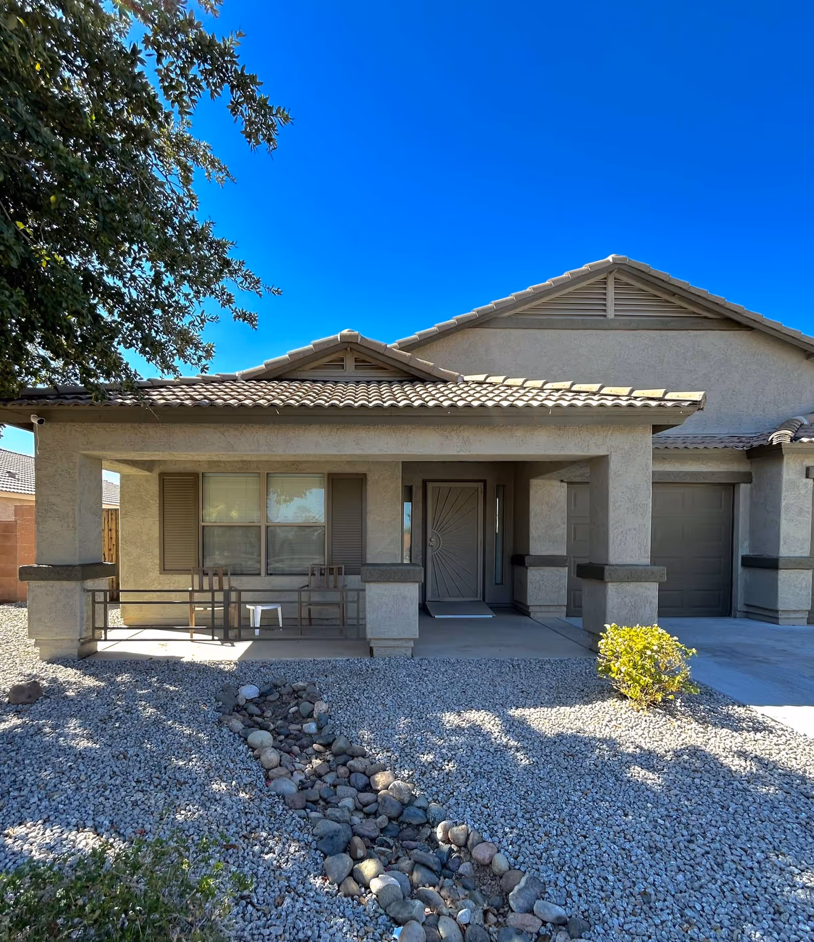 Front exterior view of a single-story house with a covered porch featuring two chairs and a small table. The house has a tiled roof, beige stucco walls, and a driveway leading to a garage. The yard is landscaped with gravel and a dry rock stream bed, with a small bush near the driveway and a tree providing shade on the left side.