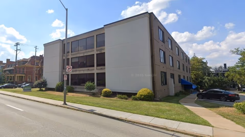 Three-story beige building with large windows along a street and a blue entrance awning.