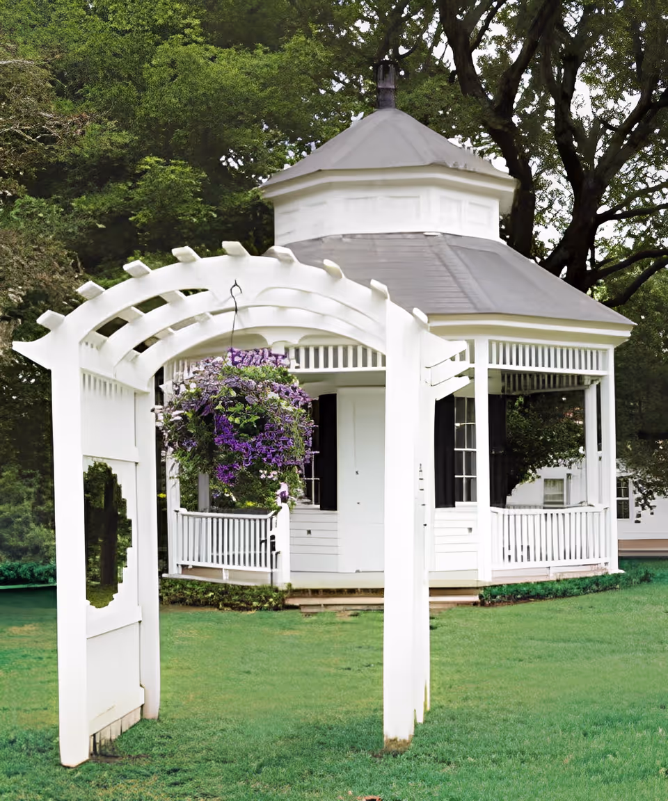 White gazebo with an arched pergola and hanging purple flowers on a green lawn surrounded by trees.