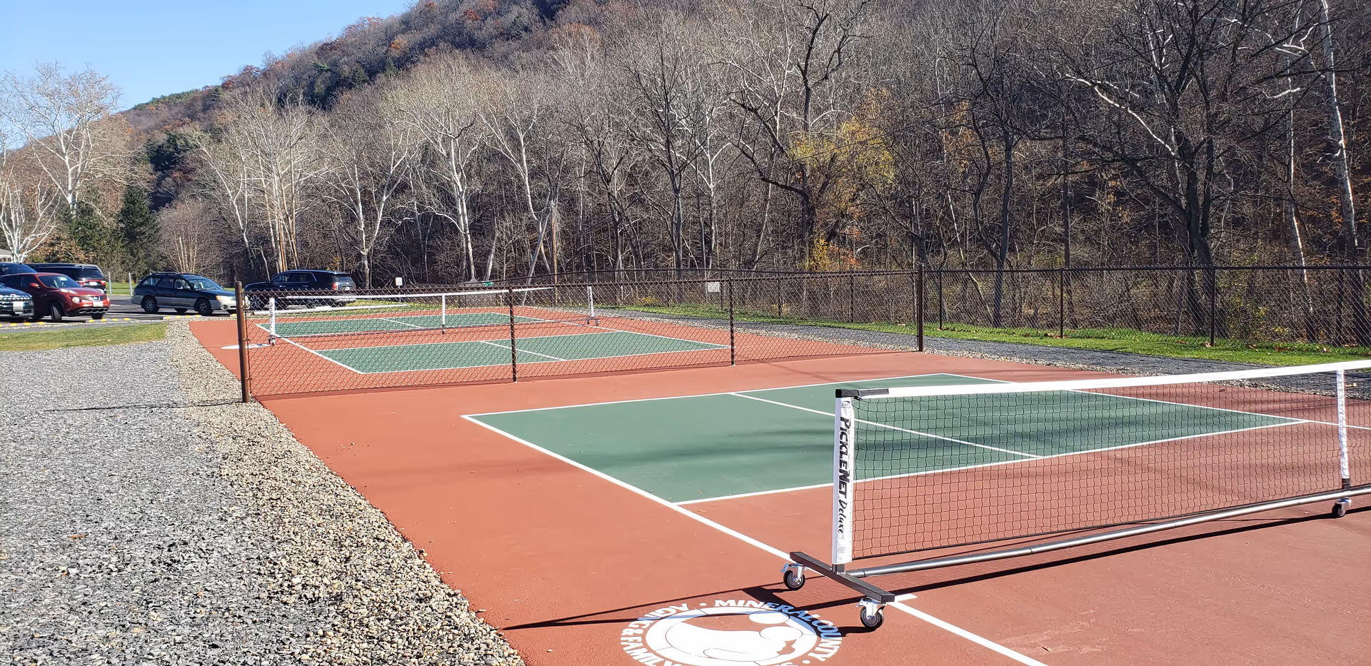 Red and green outdoor tennis court with nets, fencing, and nearby parked cars.