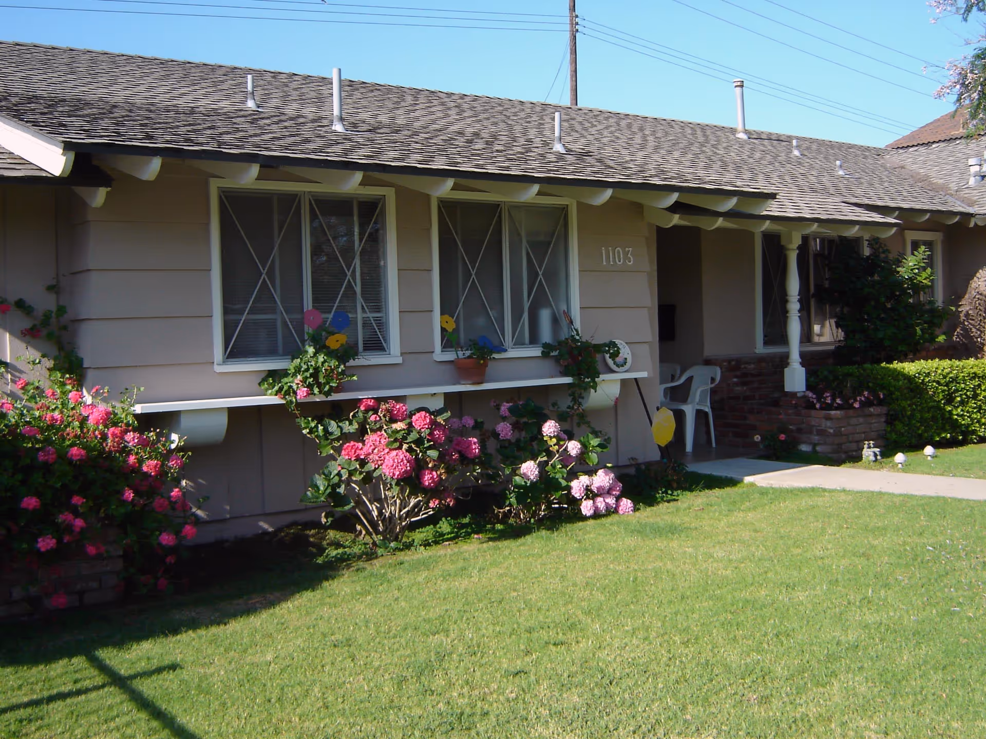 Exterior view of a single-story residential building with beige siding, two windows with white frames and decorative crossbars, a small covered porch with two white plastic chairs, and a well-maintained green lawn with blooming pink and purple flowers in front.