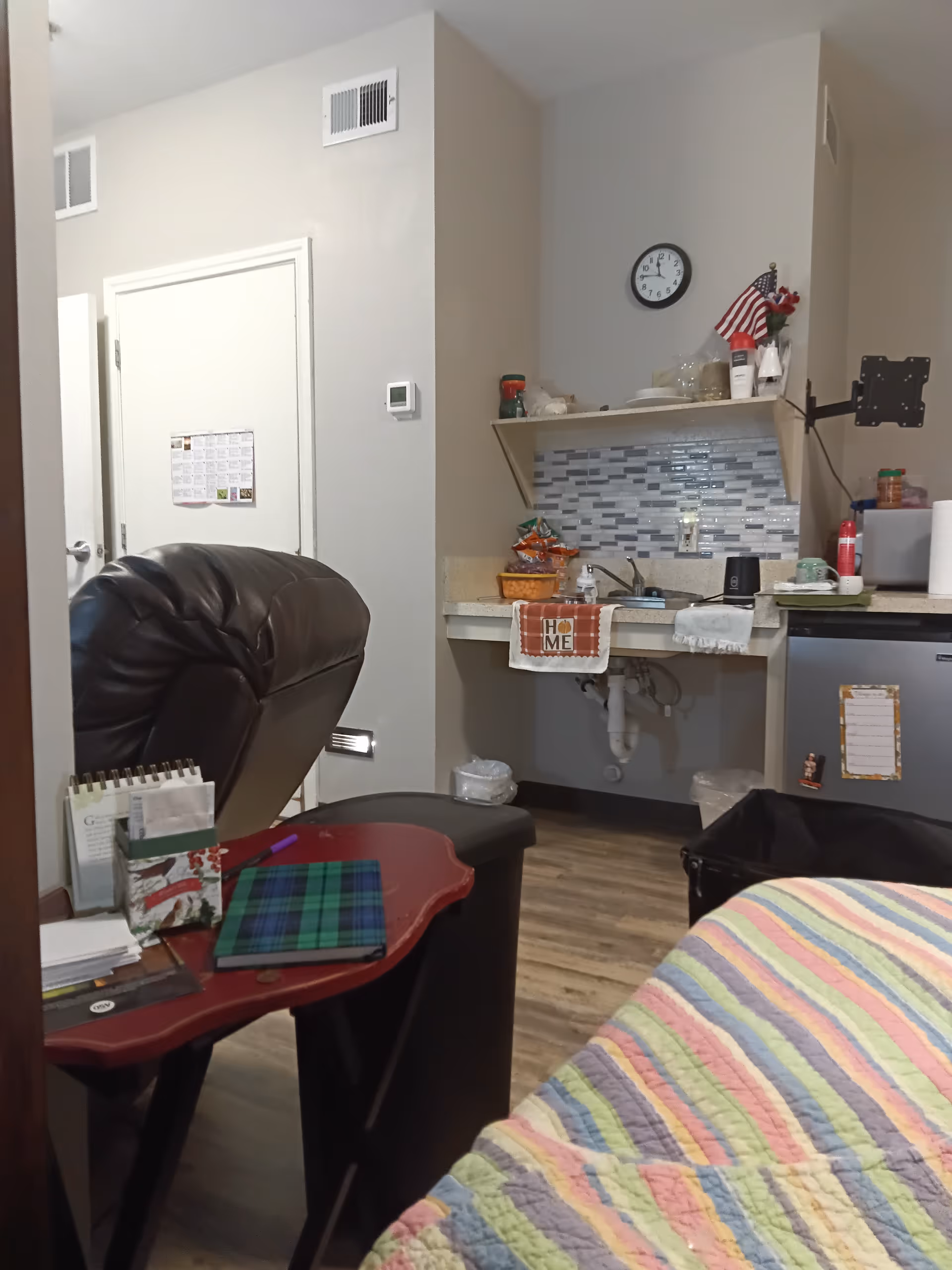 Interior living area with a small kitchenette, clock, American flag decor, and colorful quilt.