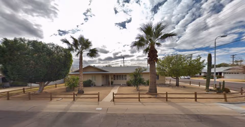 Single-story residential building with a front yard featuring palm trees, a trimmed large bush, and a wooden fence along the sidewalk under a partly cloudy sky.