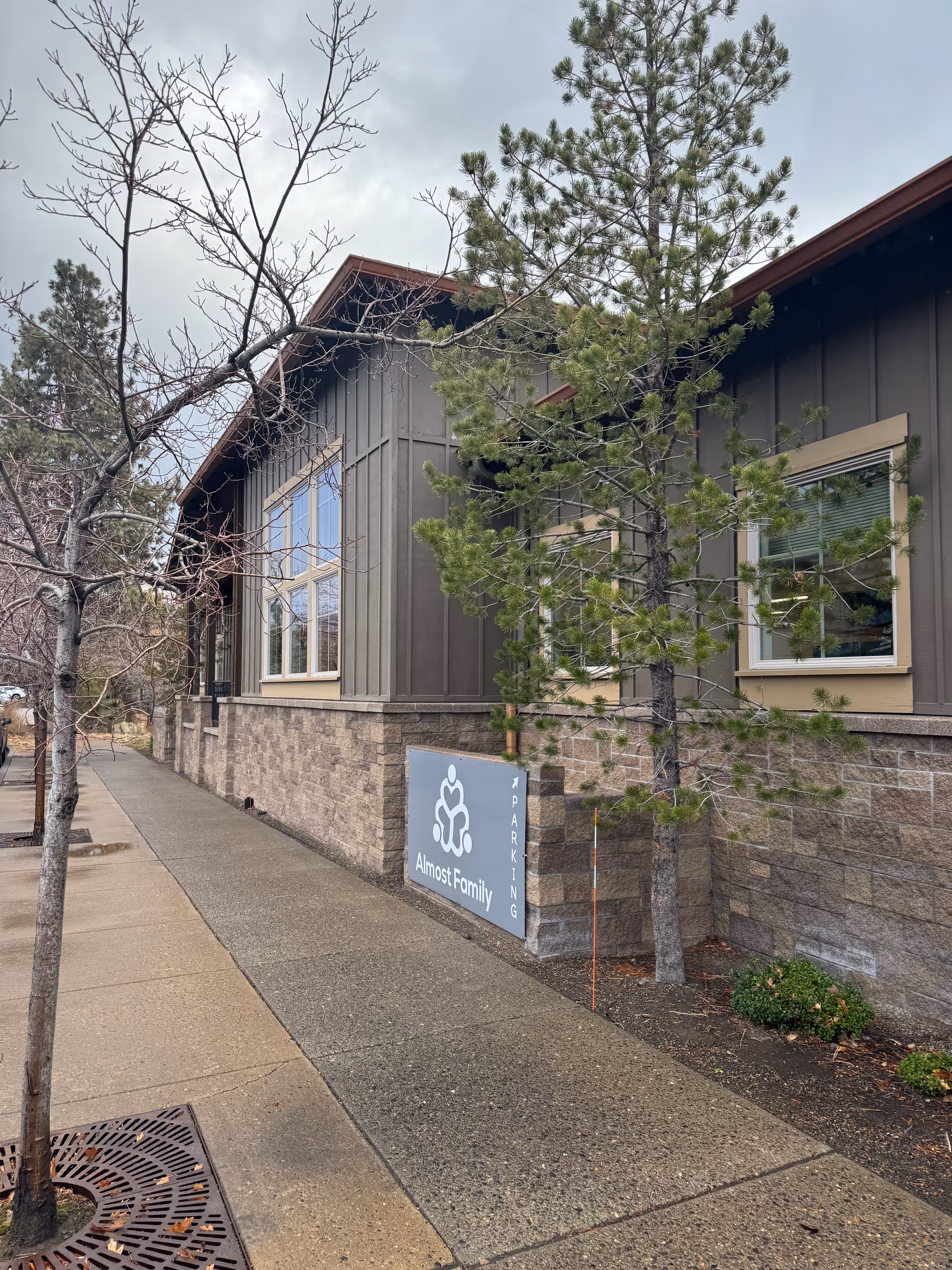 Exterior view of Almost Family facility with sidewalk, trees, and a blue parking sign.