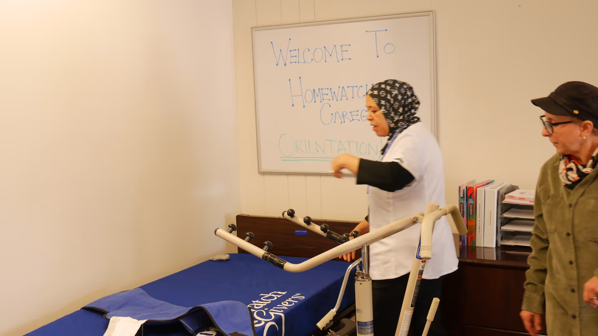Two caregivers stand by a patient lift in an orientation room.