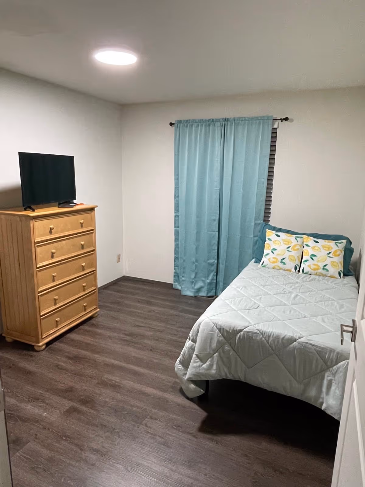 A simple bedroom with a bed, dresser, TV, and teal curtains.