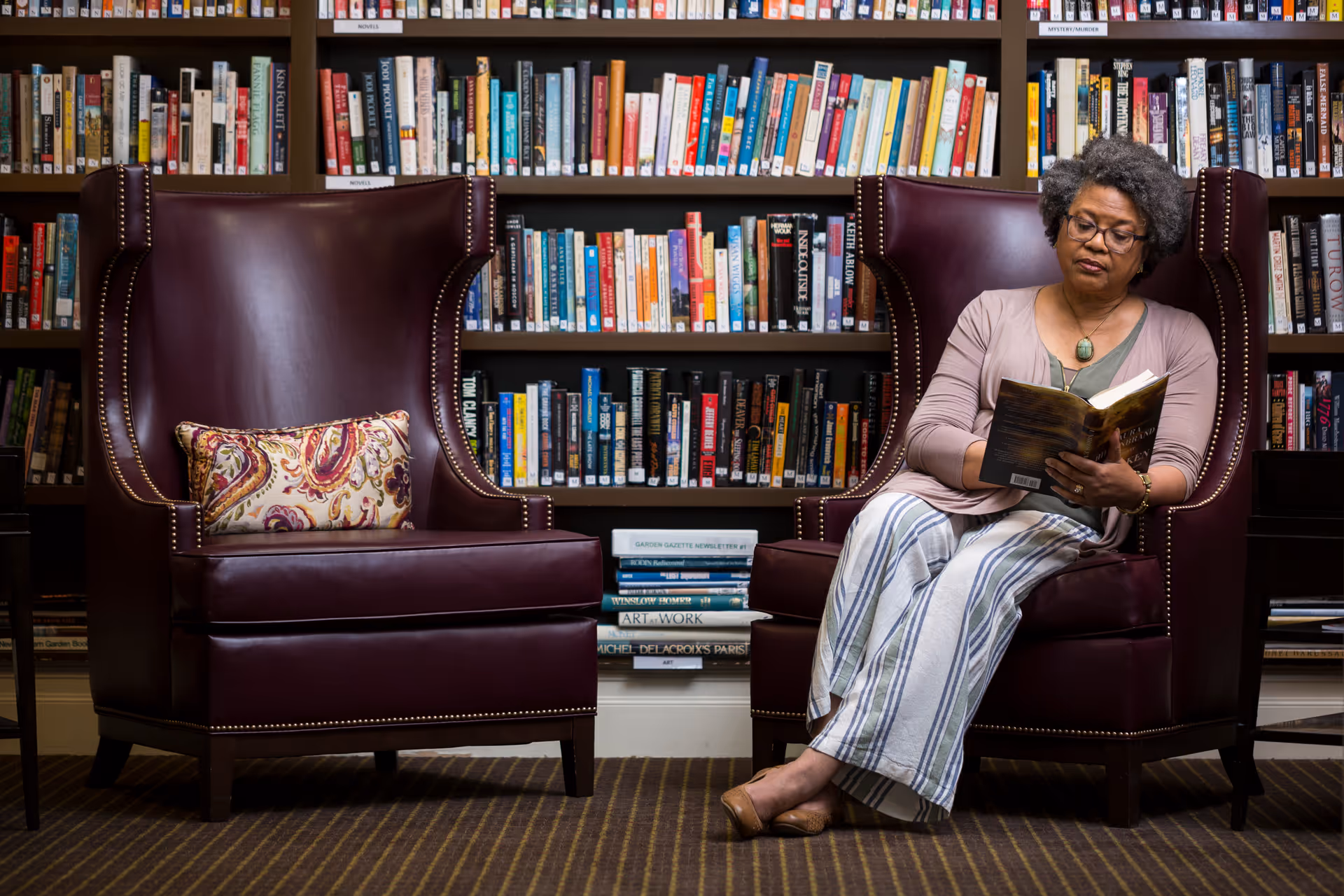 A woman with gray curly hair and glasses is sitting in a dark brown leather armchair reading a book. Another matching armchair with a decorative pillow is next to her. Behind them is a large bookshelf filled with books. The setting appears to be a quiet library or reading room.