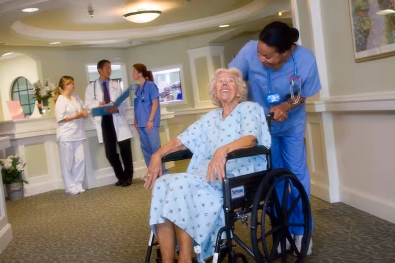 Elderly woman in a wheelchair is assisted by a nurse in a bright hospital-like hallway.