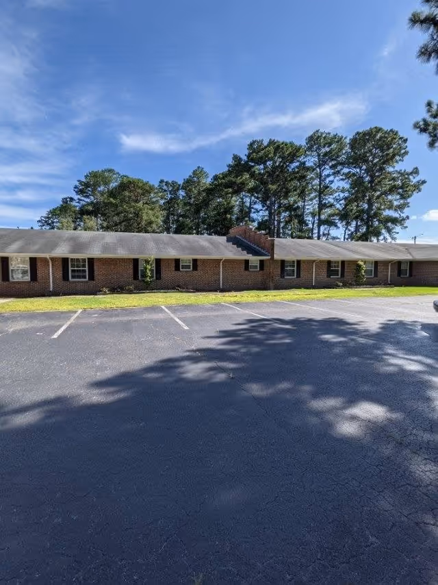Single-story brick building with a row of windows and a parking lot in front, set against tall trees and a blue sky.