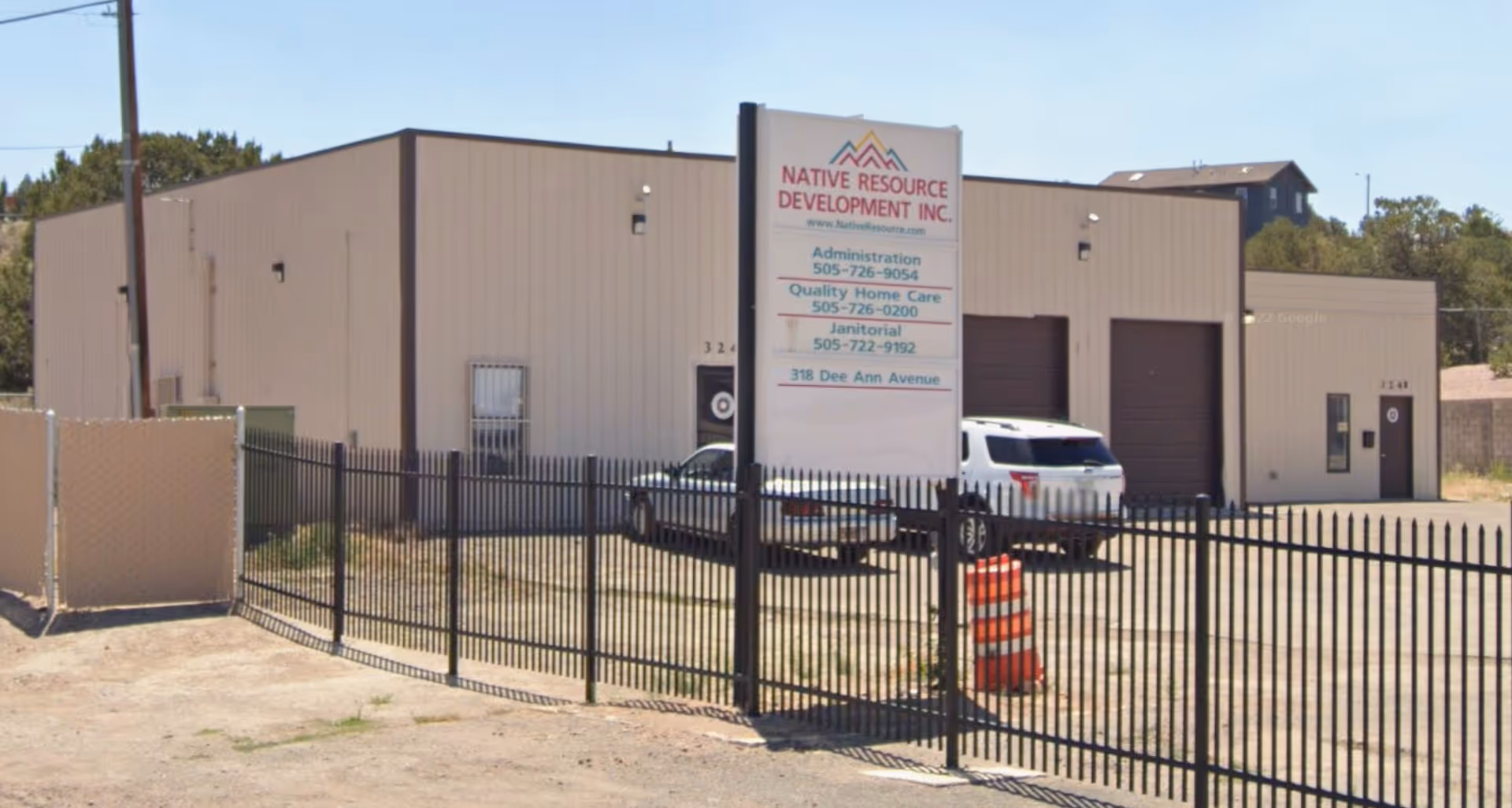 Exterior view of a beige industrial building with a sign, fence, and parked car.