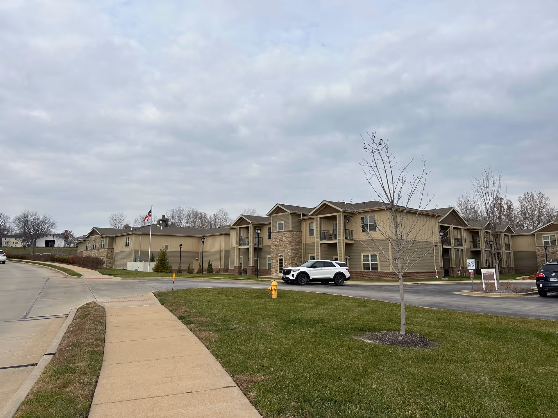 Exterior front view of a two-story senior living building with a driveway, parked cars, a sidewalk, and an American flag under a cloudy sky.