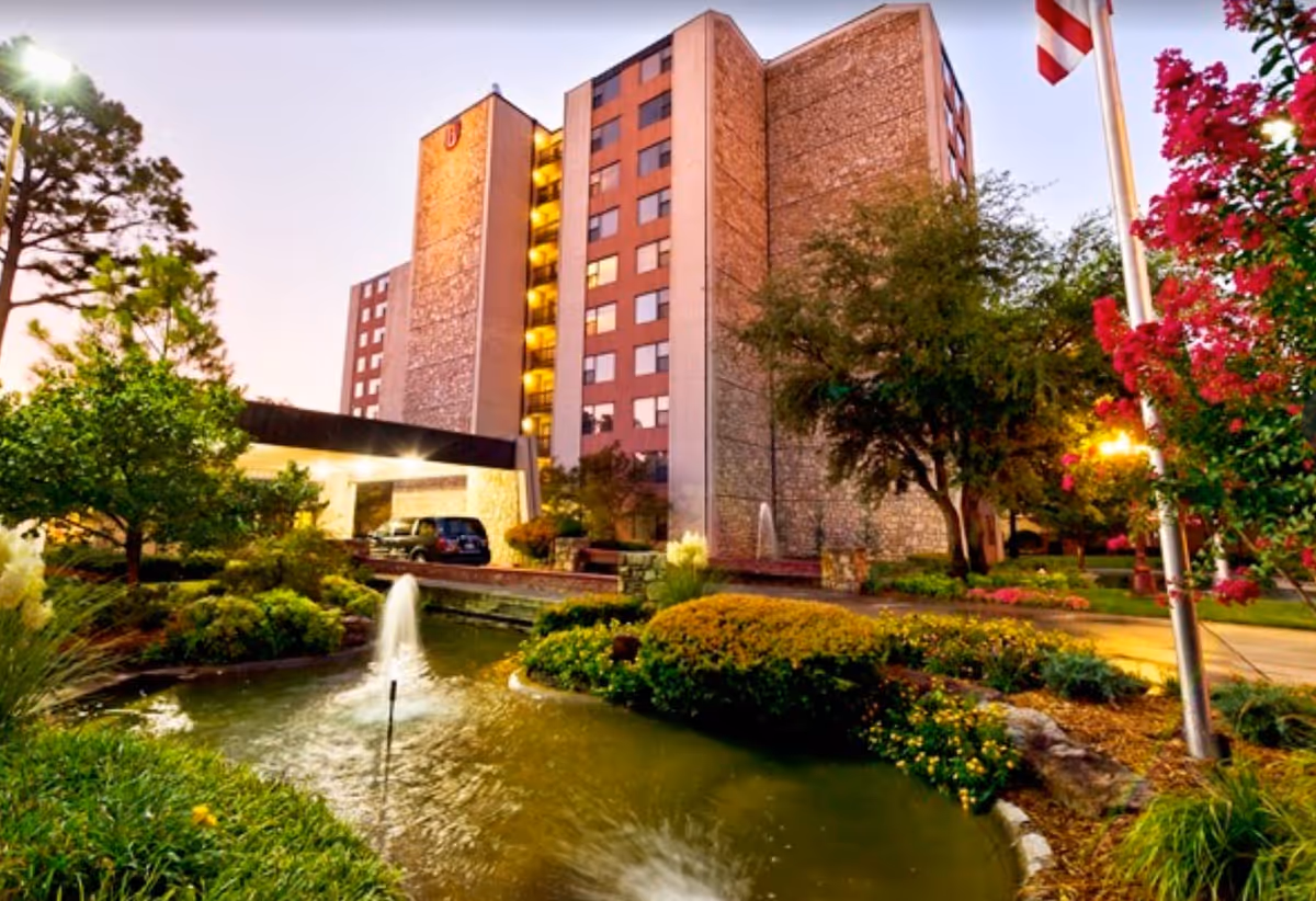 Exterior view of a multi-story senior living facility named Senior Star at Burgundy Place during dusk, featuring a well-maintained garden with a pond and fountains in the foreground, trees, flowering bushes, and an American flag on a flagpole.