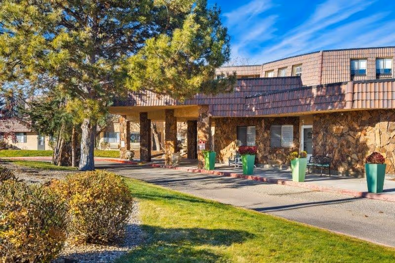 Exterior view of The Chateau de Boise senior living community building with stone facade, large windows, a covered entrance, green potted plants, trees, and well-maintained landscaping under a clear blue sky.