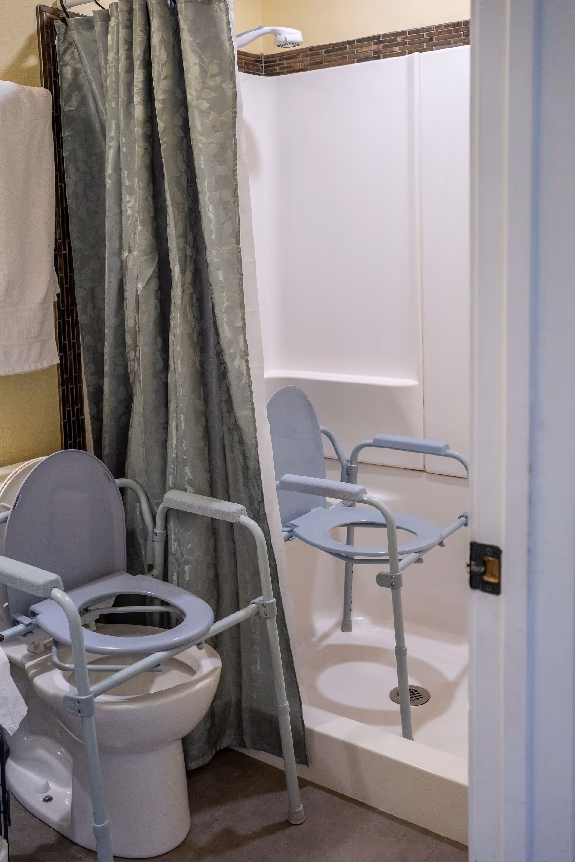 Small bathroom with a shower and two gray commode/transfer chairs, one over the toilet and one inside the shower.