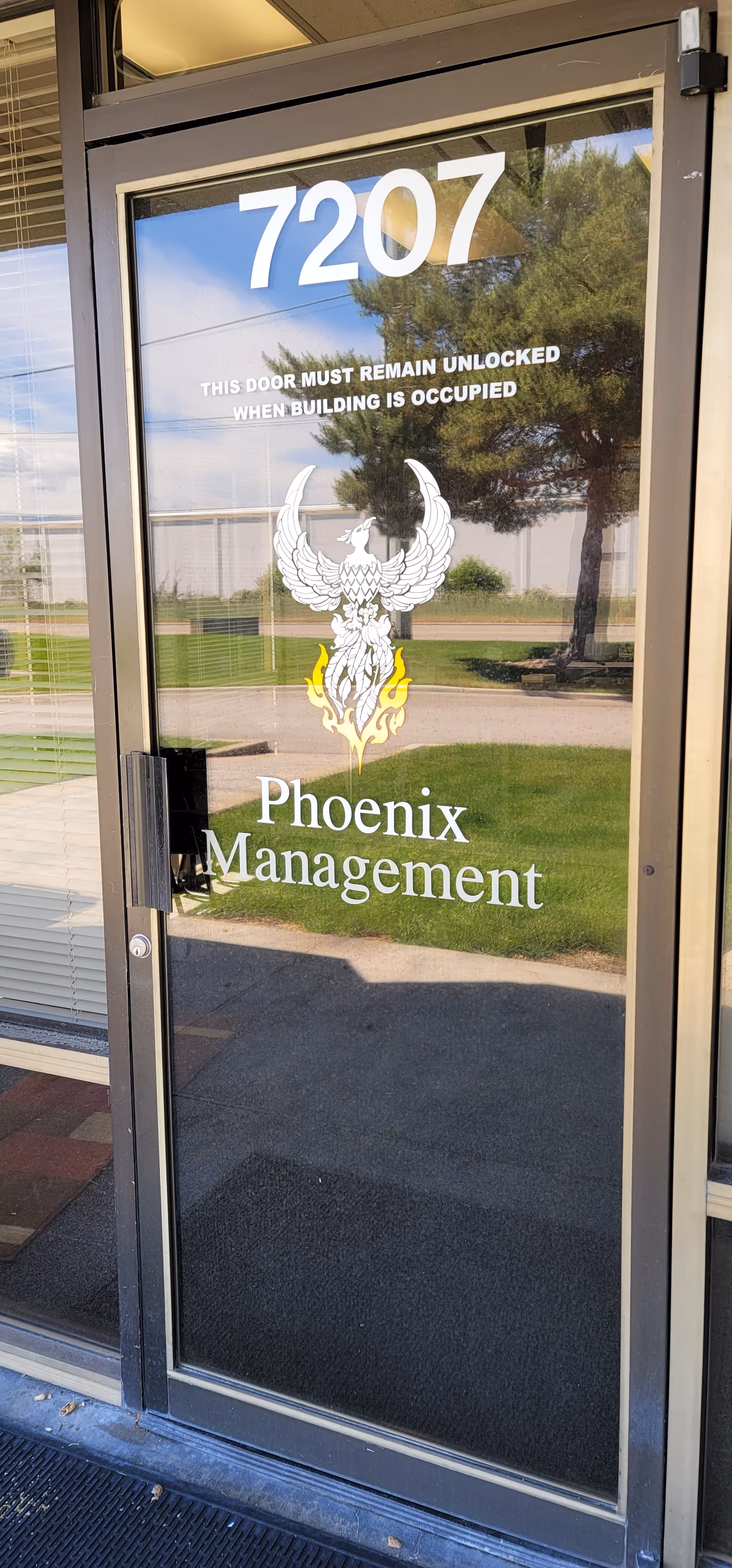 Glass entrance door with Phoenix Management logo and address 7207, reflecting trees outside.
