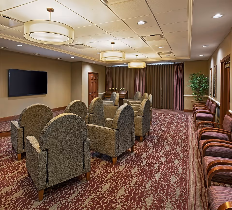A cozy common room with patterned carpet, multiple upholstered armchairs arranged in rows facing a wall-mounted flat screen TV, additional chairs lined up along the right wall, three large circular ceiling lights, and a potted plant near the back wall with closed curtains.