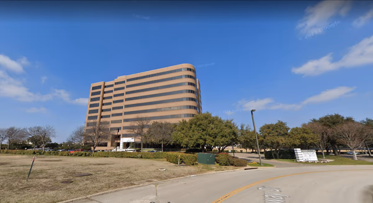 A large multi-story beige building with a curved corner stands beside a road under blue sky.
