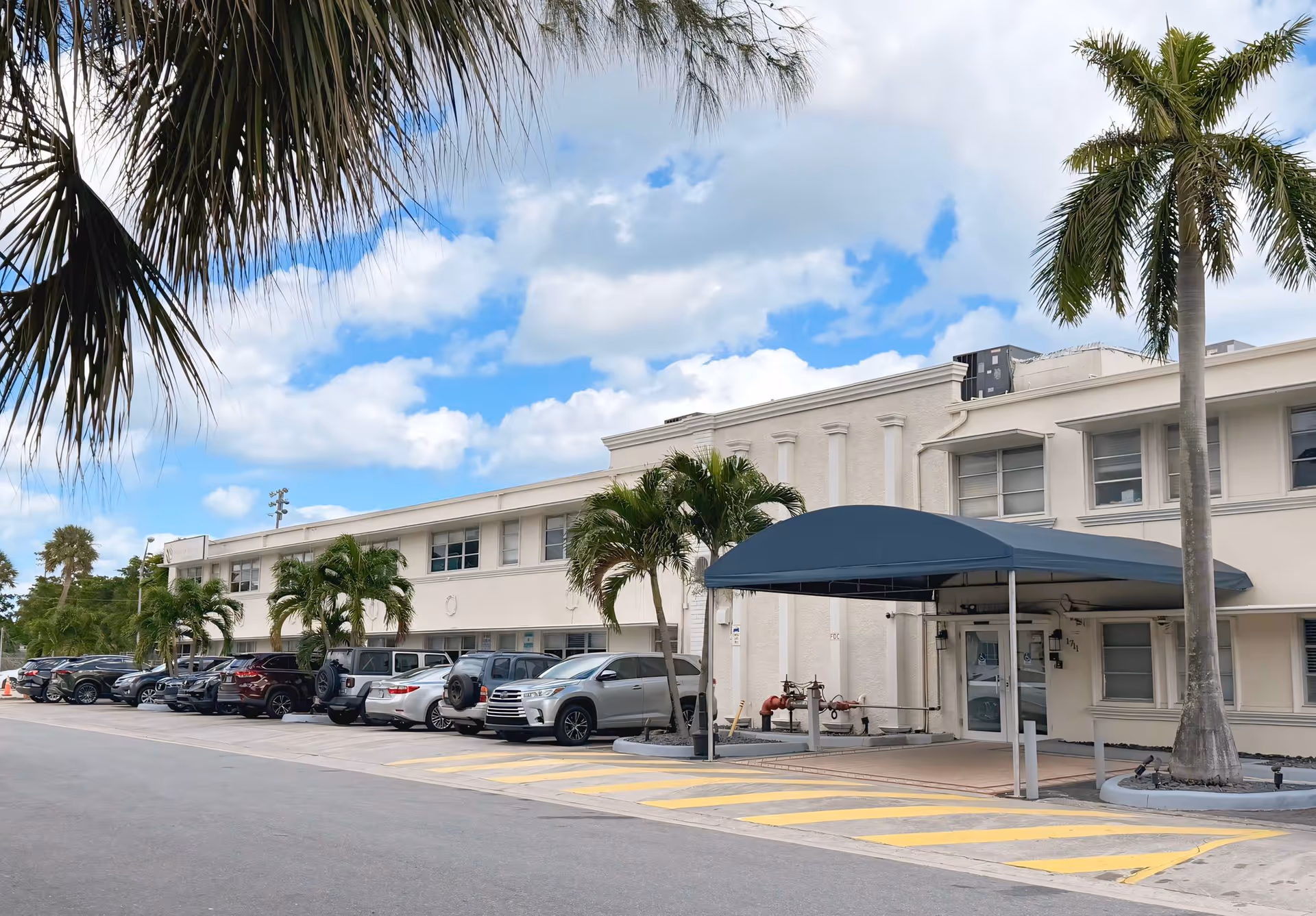 Exterior view of a two-story white building with a covered entrance and several parked cars along the side. Palm trees are visible near the building and in the foreground, under a partly cloudy blue sky.
