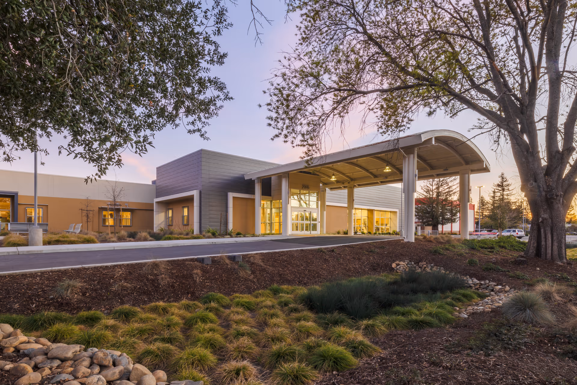 Senior living facility exterior with glass doors, canopy, and landscaped surroundings at sunset.