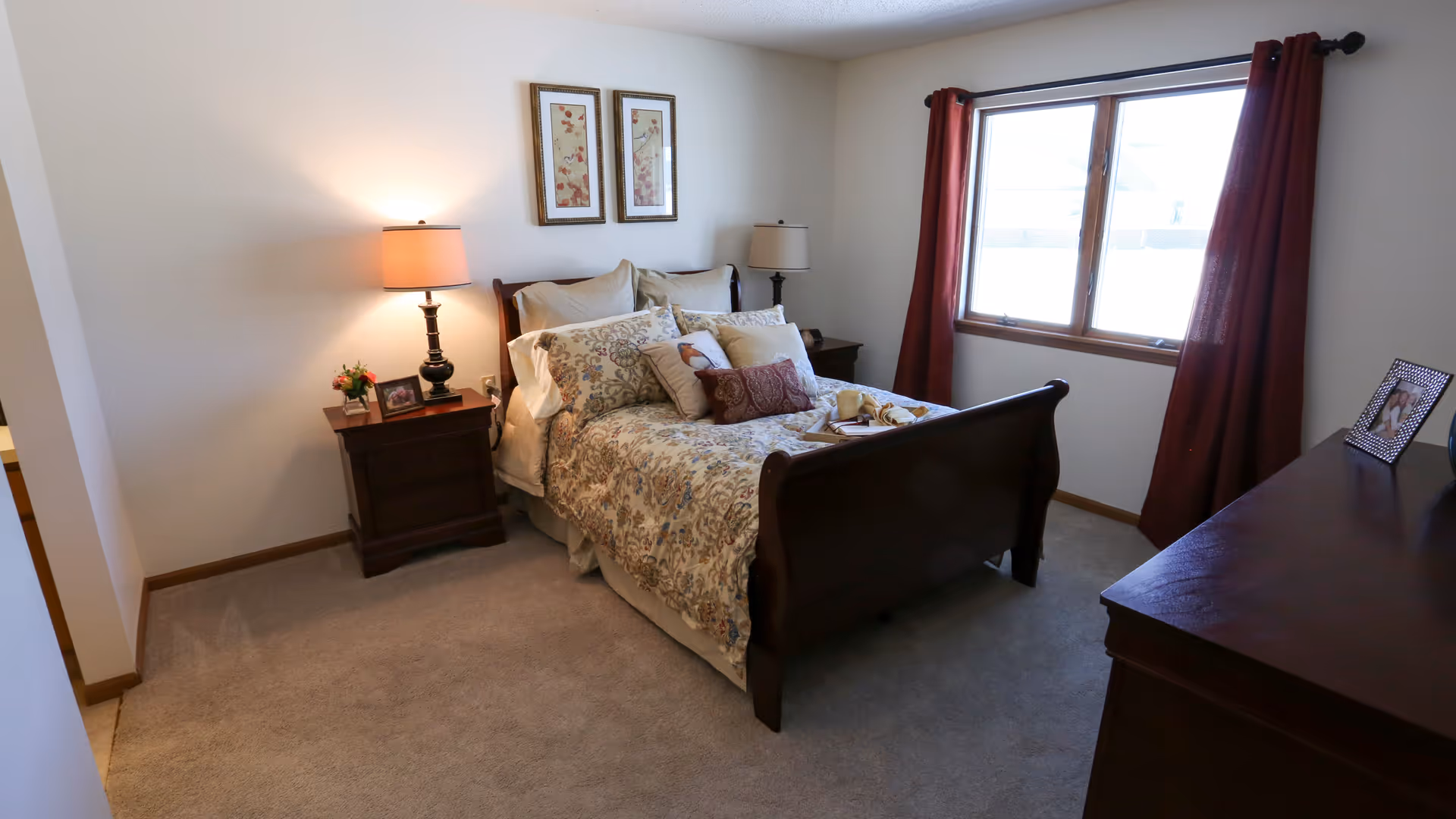 Cozy bedroom with a wooden bed, matching nightstands, lamps, and a window with red curtains.