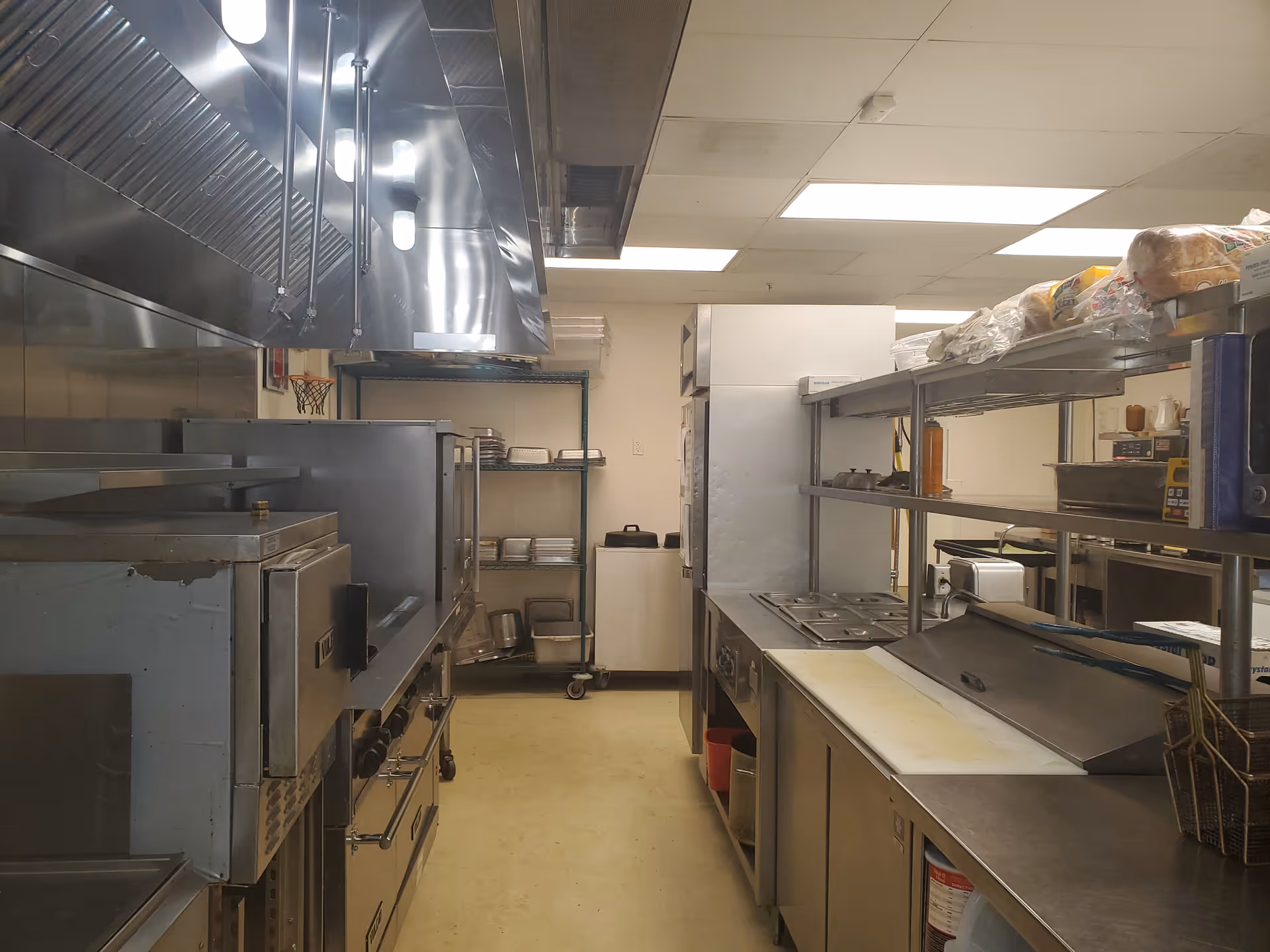 Commercial stainless-steel kitchen with ovens, shelves, and overhead venting in a facility.