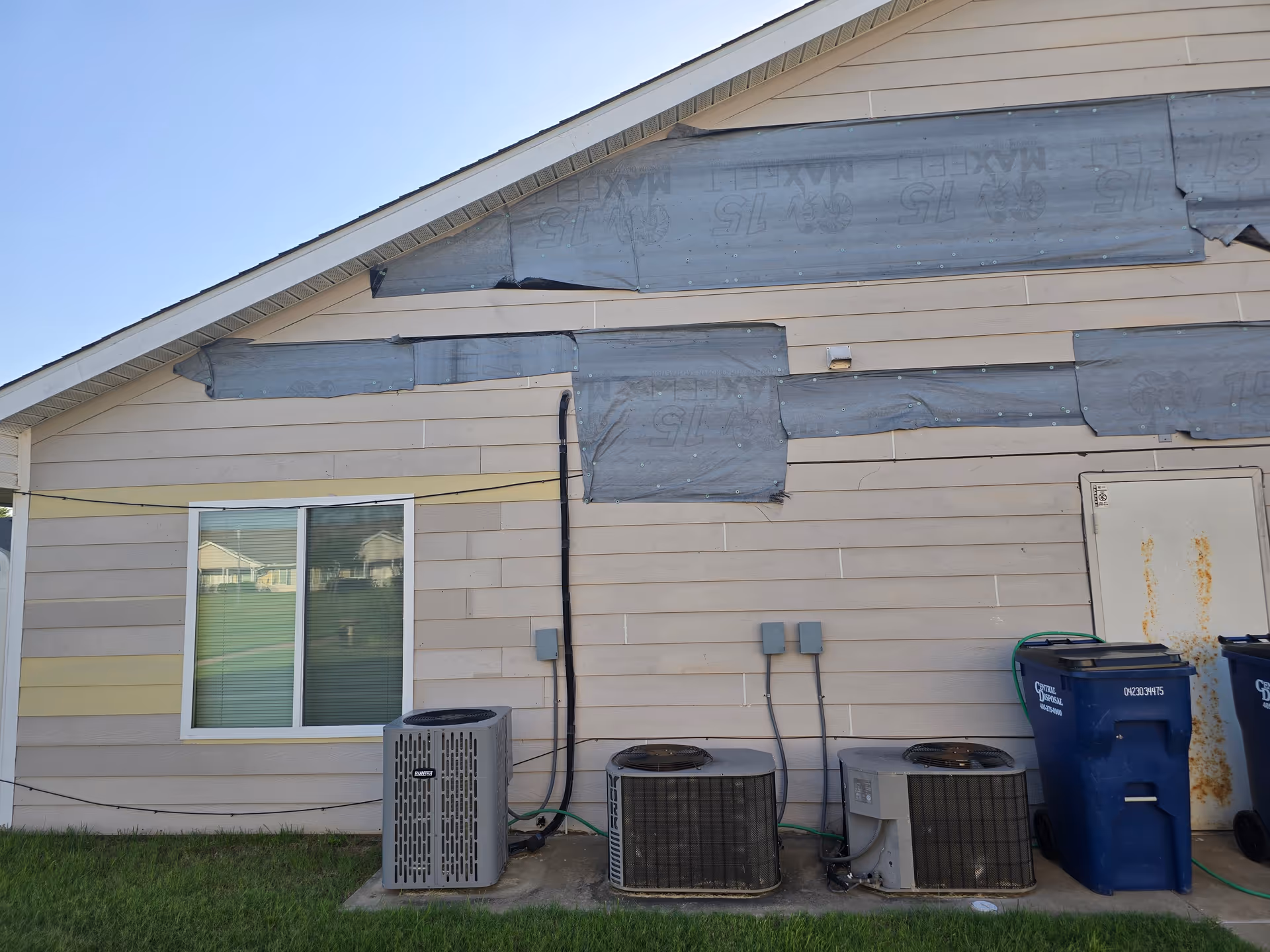 Side exterior of a beige apartment building with air conditioning units and trash bins.