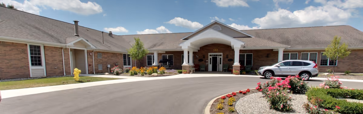 Single-story brick senior living facility entrance with a covered portico, circular driveway, landscaped flowerbeds and a parked car.