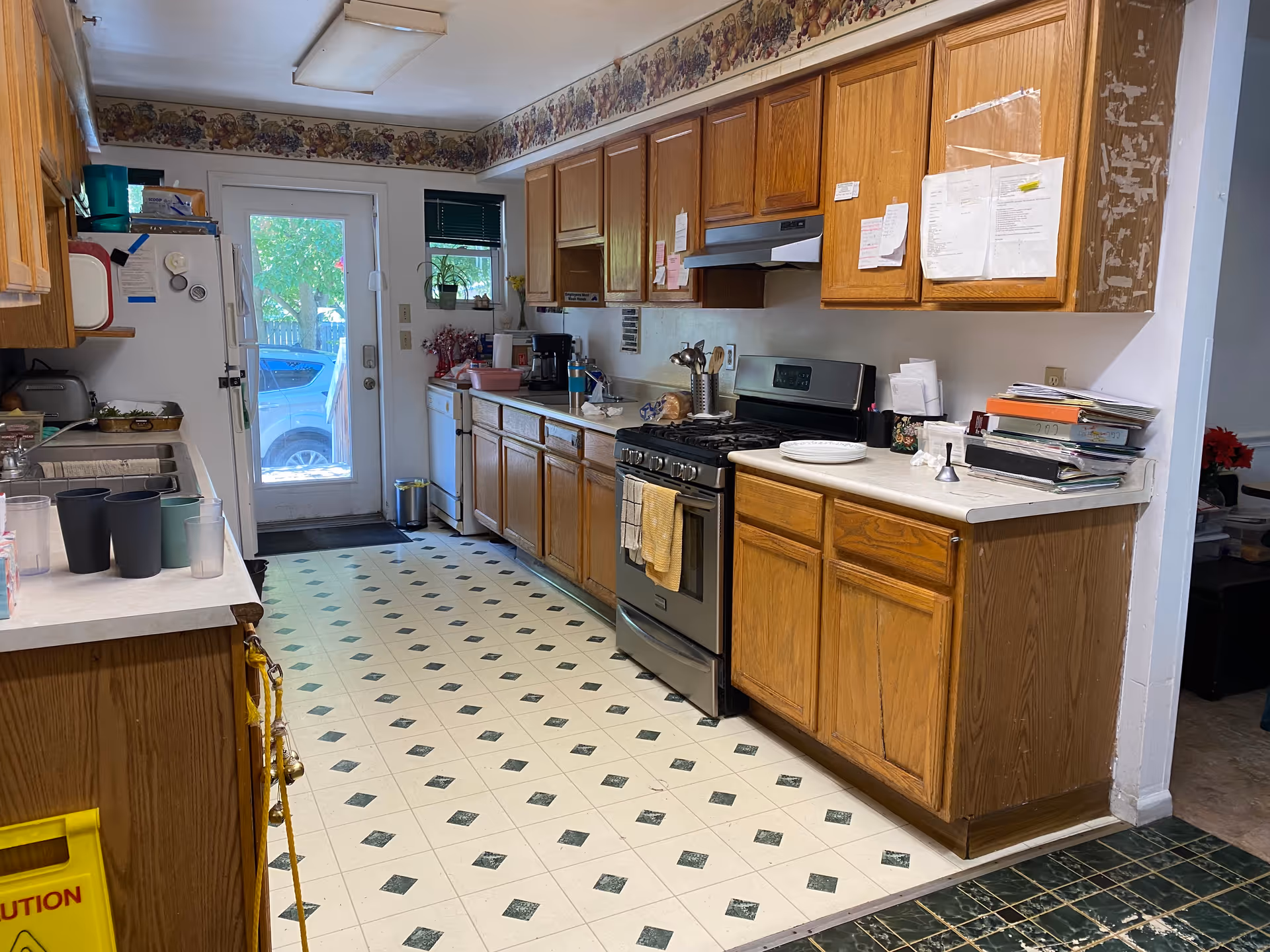 Long kitchen with wooden cabinets, appliances, and a door to outside; cluttered counters.