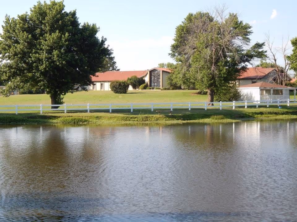Franciscan Villa exterior with red-roofed buildings, white fence, green lawn, and pond foreground.