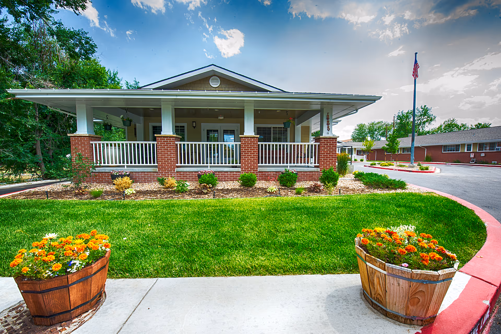 Front exterior view of a single-story building with a covered porch supported by white columns and red brick bases. The building is surrounded by a well-maintained lawn with flower beds and two wooden planters filled with orange and white flowers. A flagpole with an American flag is visible on the right side near the driveway under a partly cloudy sky.