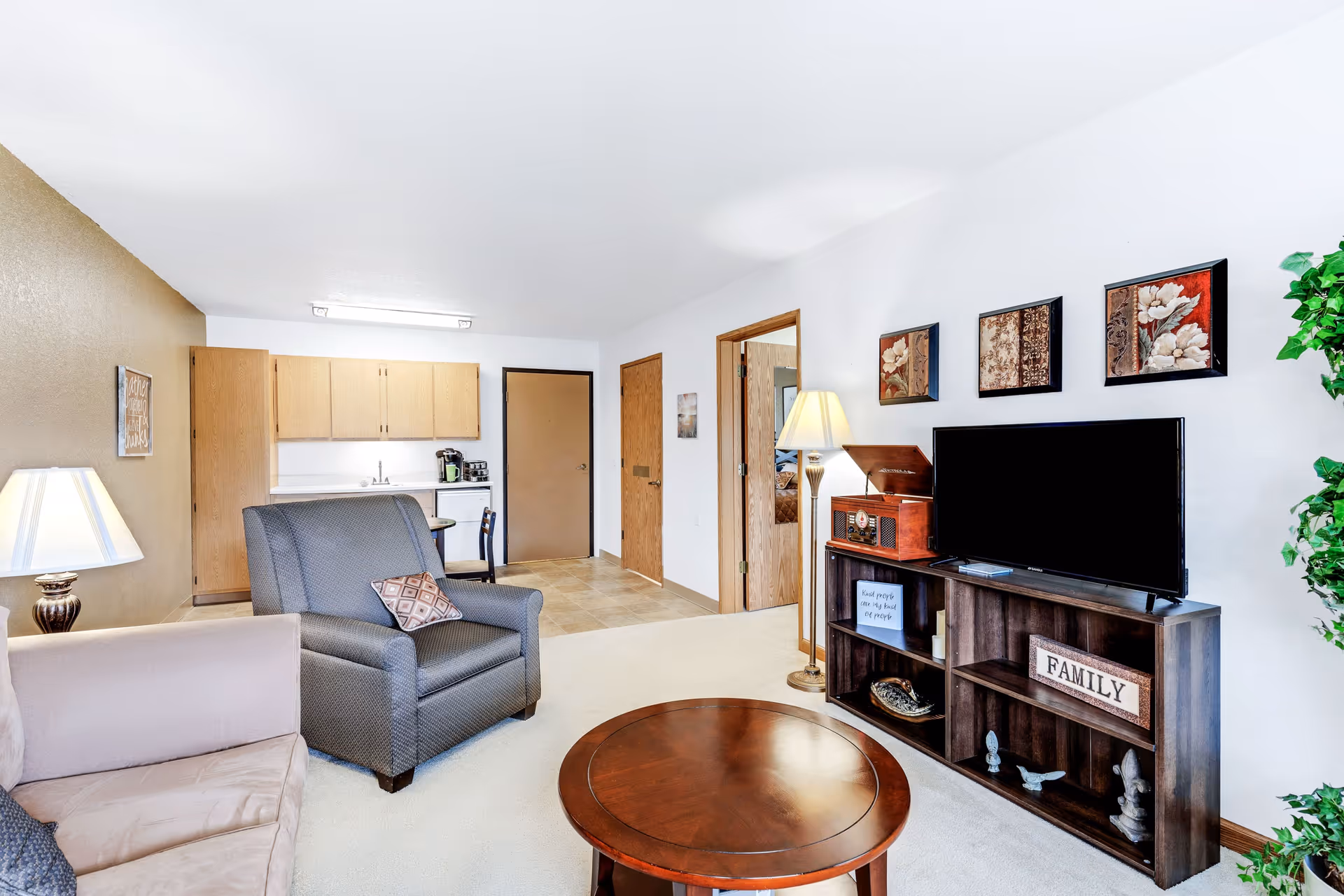 A cozy living room area in a senior living facility with a beige couch, a gray armchair, and a round wooden coffee table. There is a wooden TV stand with a flat-screen TV, decorative items, and a vintage-style record player. The walls are white with three framed floral artworks above the TV. A floor lamp stands next to the TV stand, and a green plant is visible on the right. In the background, there is a small kitchenette with wooden cabinets, a sink, a coffee maker, and a small dining table with chairs. Two wooden doors are visible, one open leading to another room.