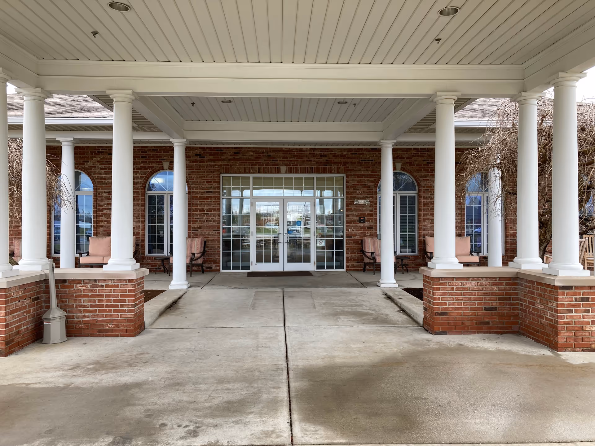 Covered front entrance of a brick building with white columns, outdoor chairs, and glass double doors.