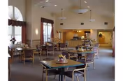 Bright dining room with multiple tables, chairs, centerpieces, and a kitchen area in the background.