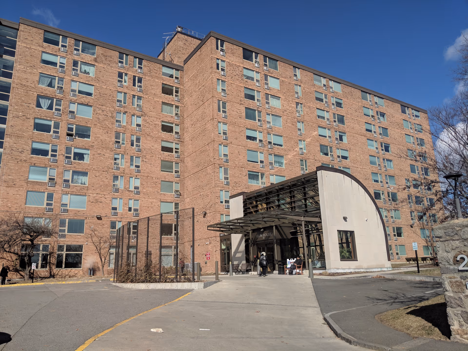 Exterior view of a large multi-story brick building under a clear blue sky. The building has many windows and a modern entrance with a glass canopy. There is a paved driveway leading up to the entrance and some trees and landscaping around the building.