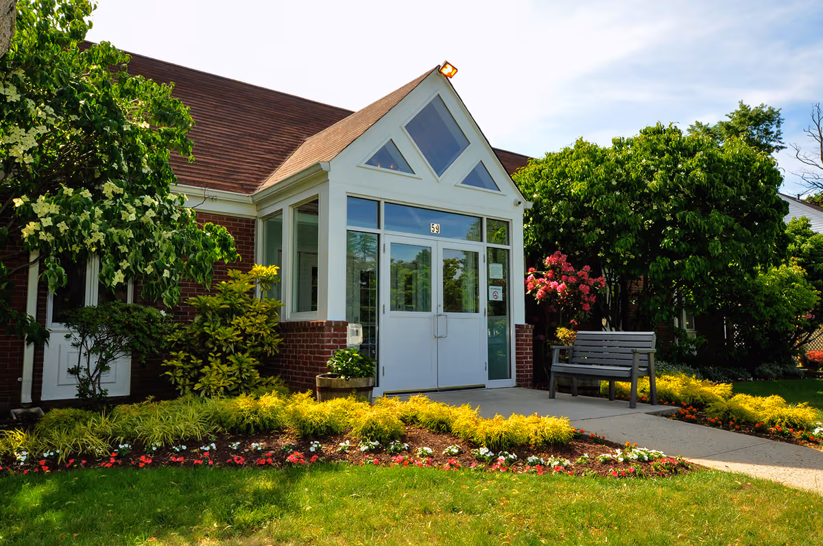 Front entrance of a brick building with glass double doors, landscaped flower beds and a bench.
