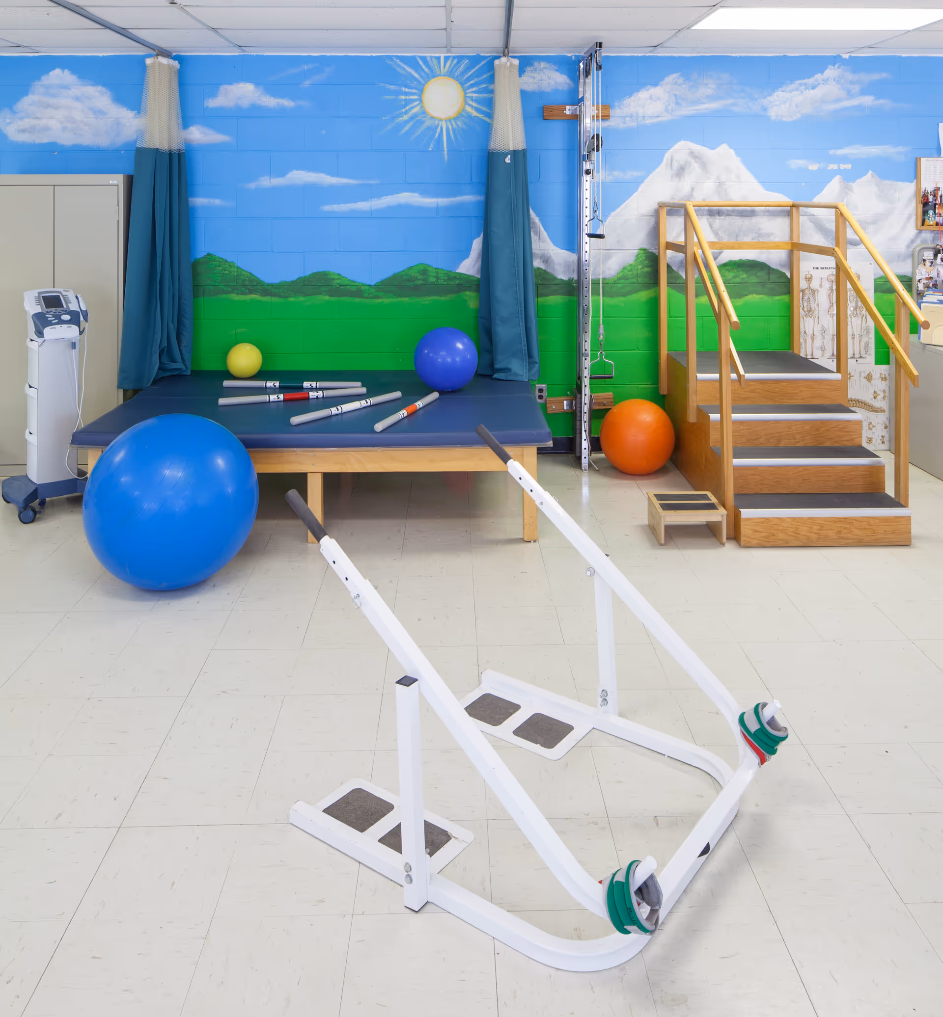 An indoor rehabilitation gym with exercise equipment, blue mural, and wooden staircase in a senior facility.