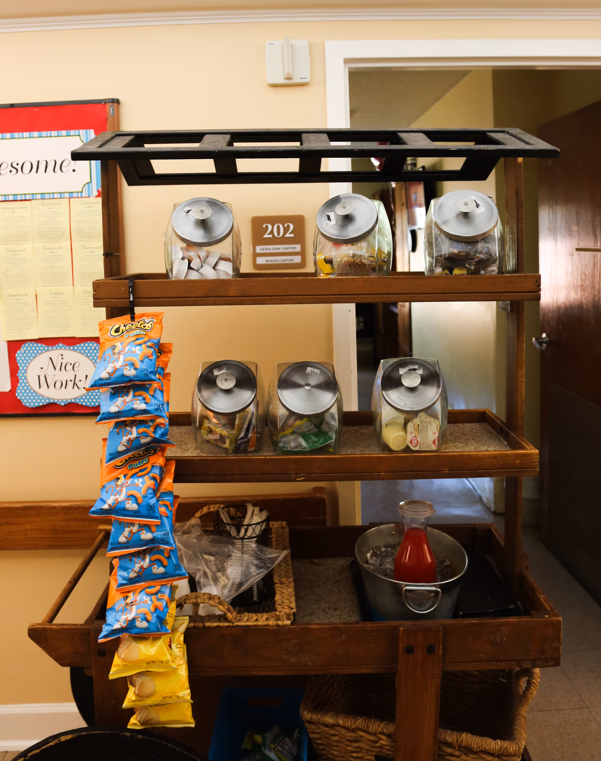 A wooden snack cart with three shelves holding various snack jars and bags of chips hanging on the side. The top shelf has three glass jars with metal lids containing snacks. The middle shelf has three more jars with snacks. The bottom shelf has a metal bucket with a bottle of red juice on ice, a basket with napkins, and other items. Behind the cart is a beige wall with a door labeled 202 and a bulletin board with a 'Nice Work!' sign.