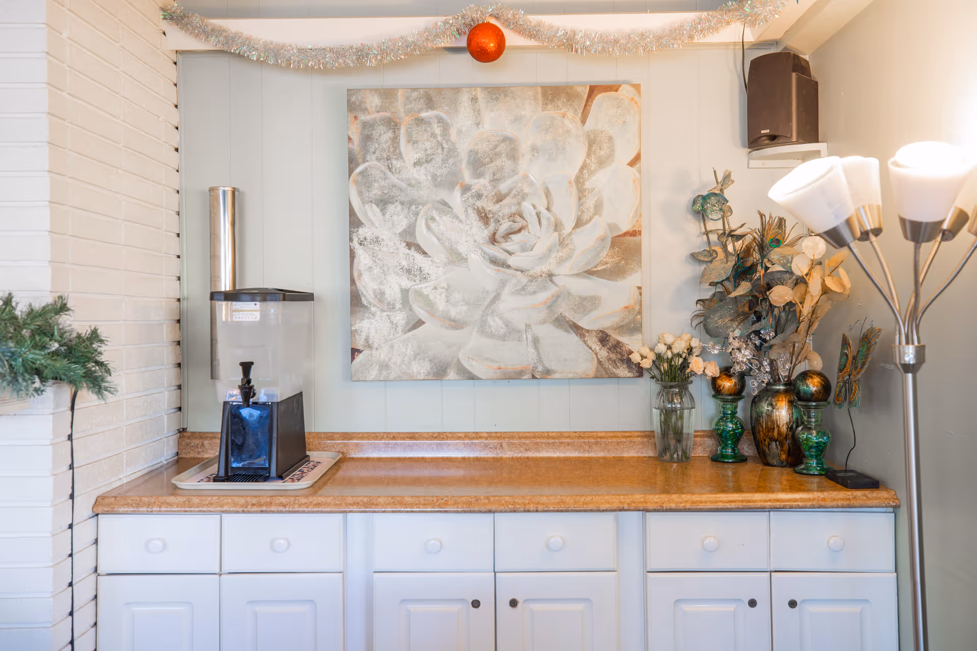 Countertop area with a water dispenser, decorative vases and flowers beneath a large floral painting and a standing lamp.