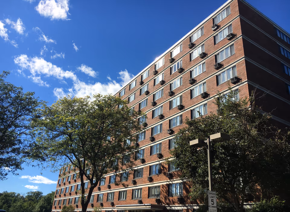 Low-angle view of a multi-story brick residential building with trees in front under a blue sky.