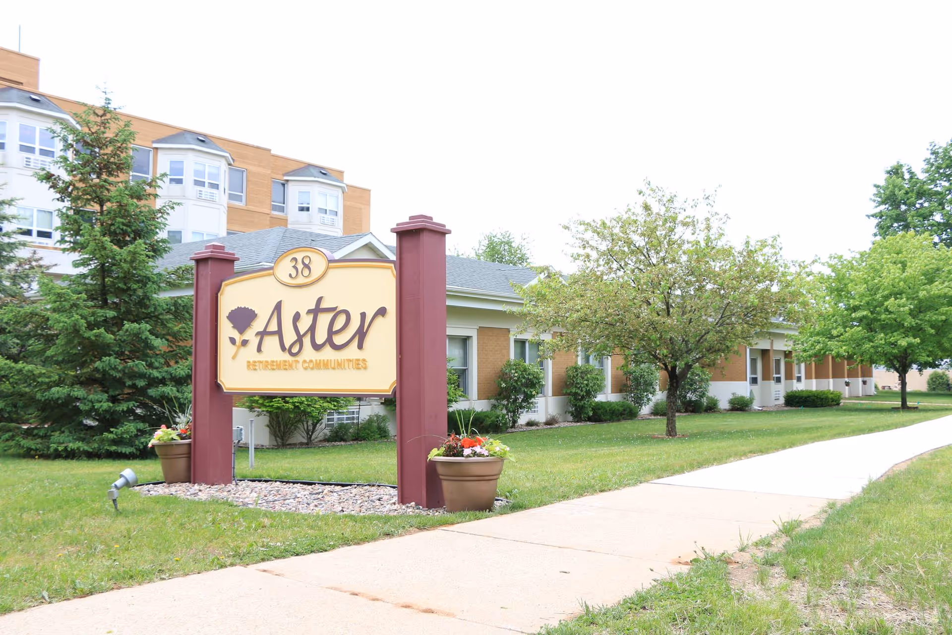 Sign reading 'Aster Retirement Communities' stands on a lawn in front of a low-rise assisted living building with trees and a sidewalk.