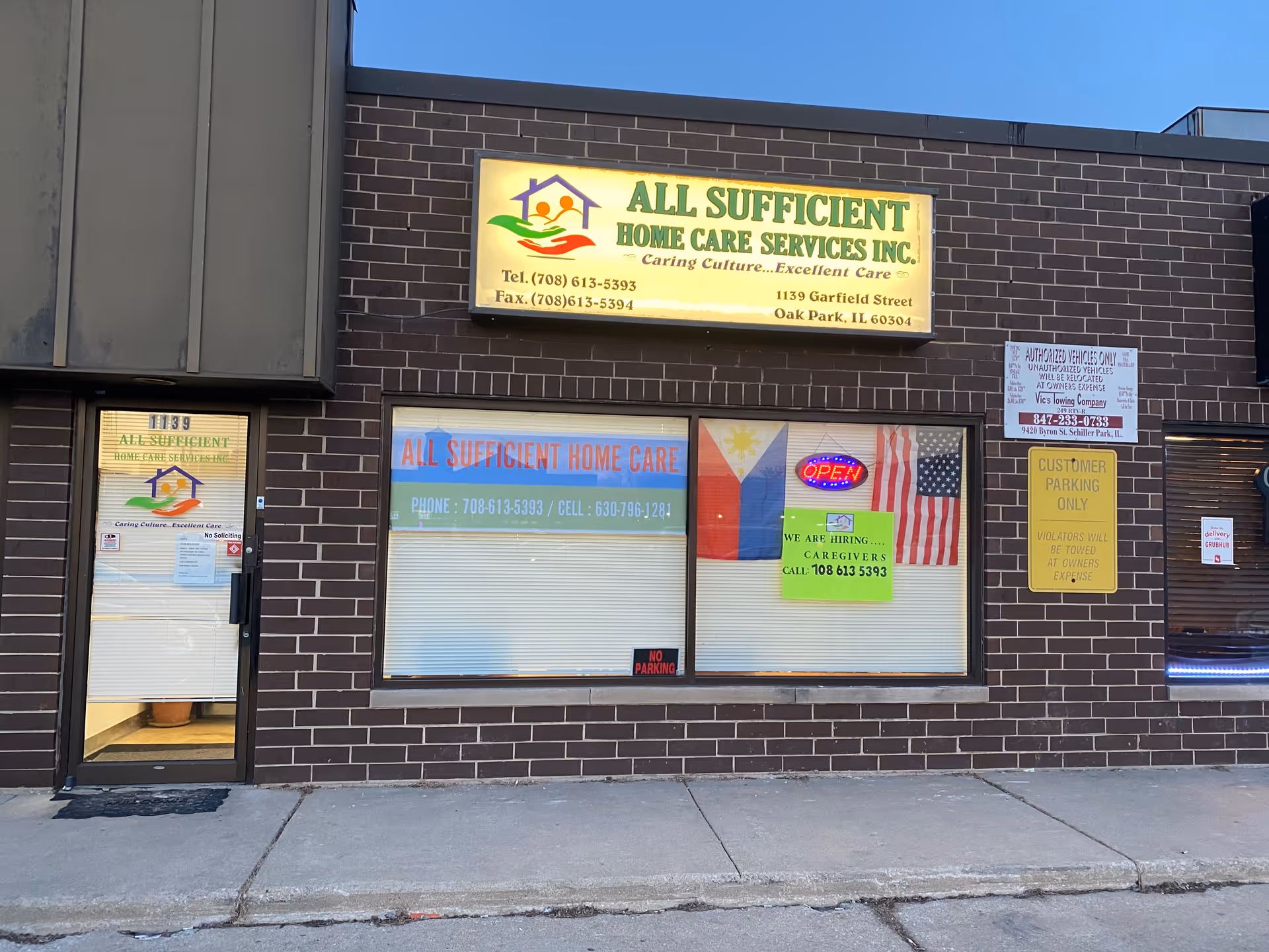 Brick storefront with All Sufficient Home Care Services sign and windows displaying hiring notice and door.