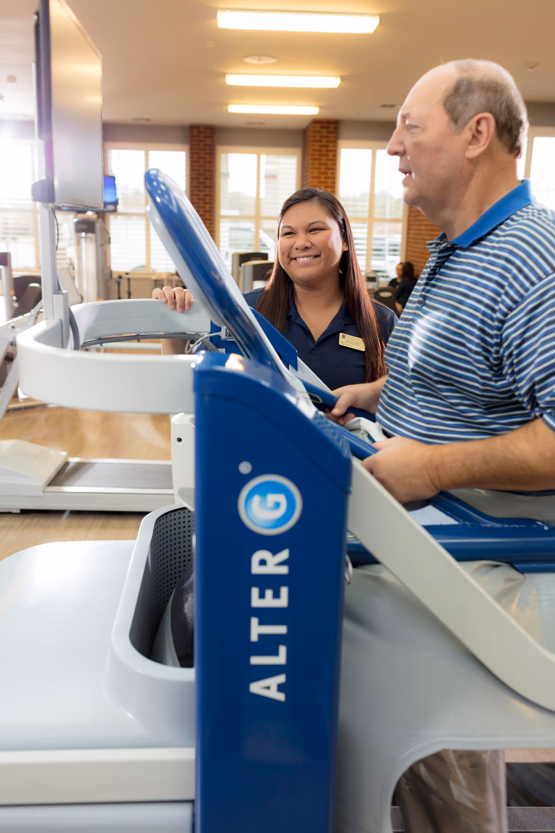 A man using an AlterG anti-gravity treadmill in a rehabilitation center while a female staff member smiles and assists him. The room has large windows with natural light and exercise equipment in the background.