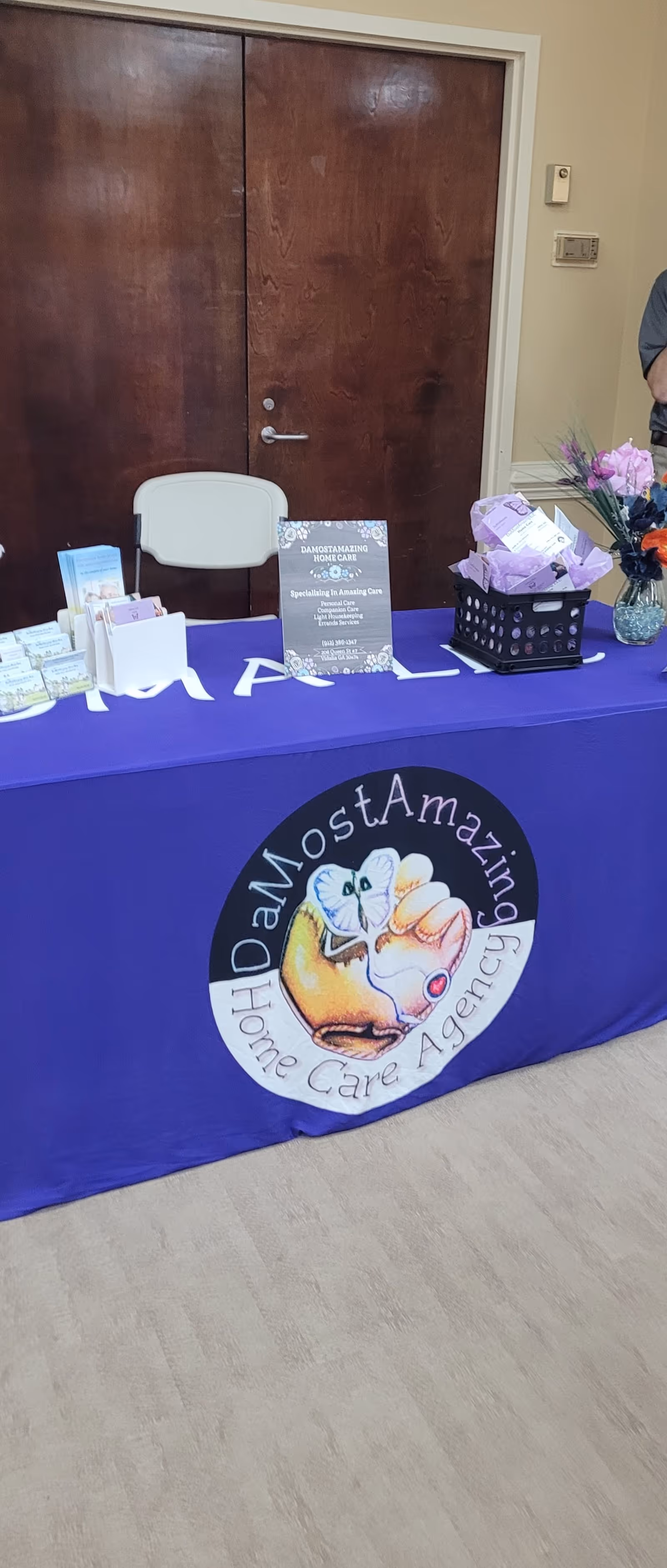 Purple tablecloth display with brochures and DaMostAmazing Home Care Agency logo indoors.