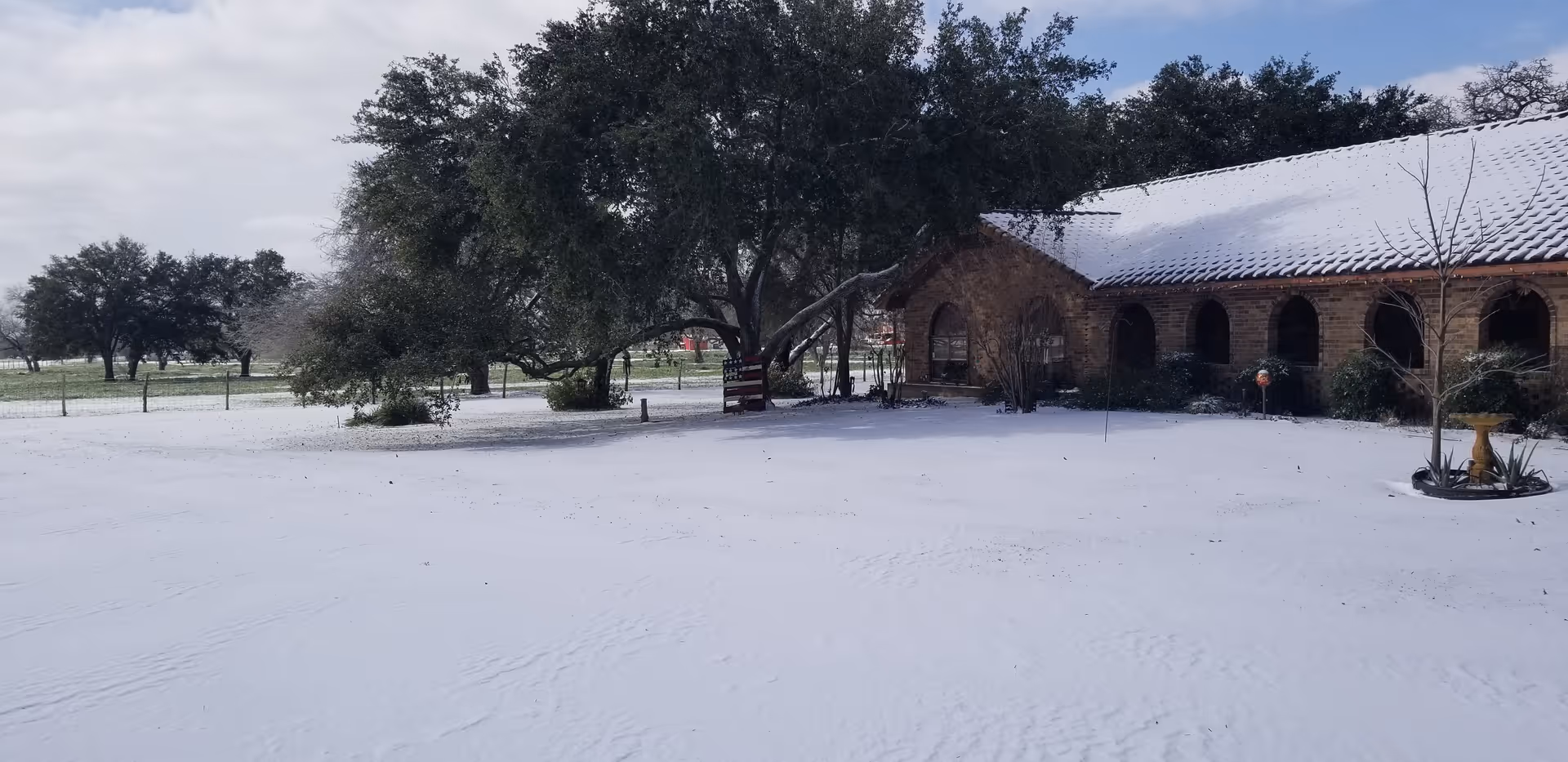A snow-covered yard with a large tree and a brick building with arched windows and a snow-covered roof. The scene is outdoors with more trees and a fence in the background under a cloudy sky.